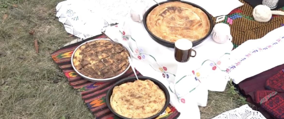 A display of traditional Serbian dishes served outdoors on a colorful woven mat. Three round trays feature different types of pies and baked goods, with a ceramic mug and decorative embroidered cloth adding to the rustic presentation. 