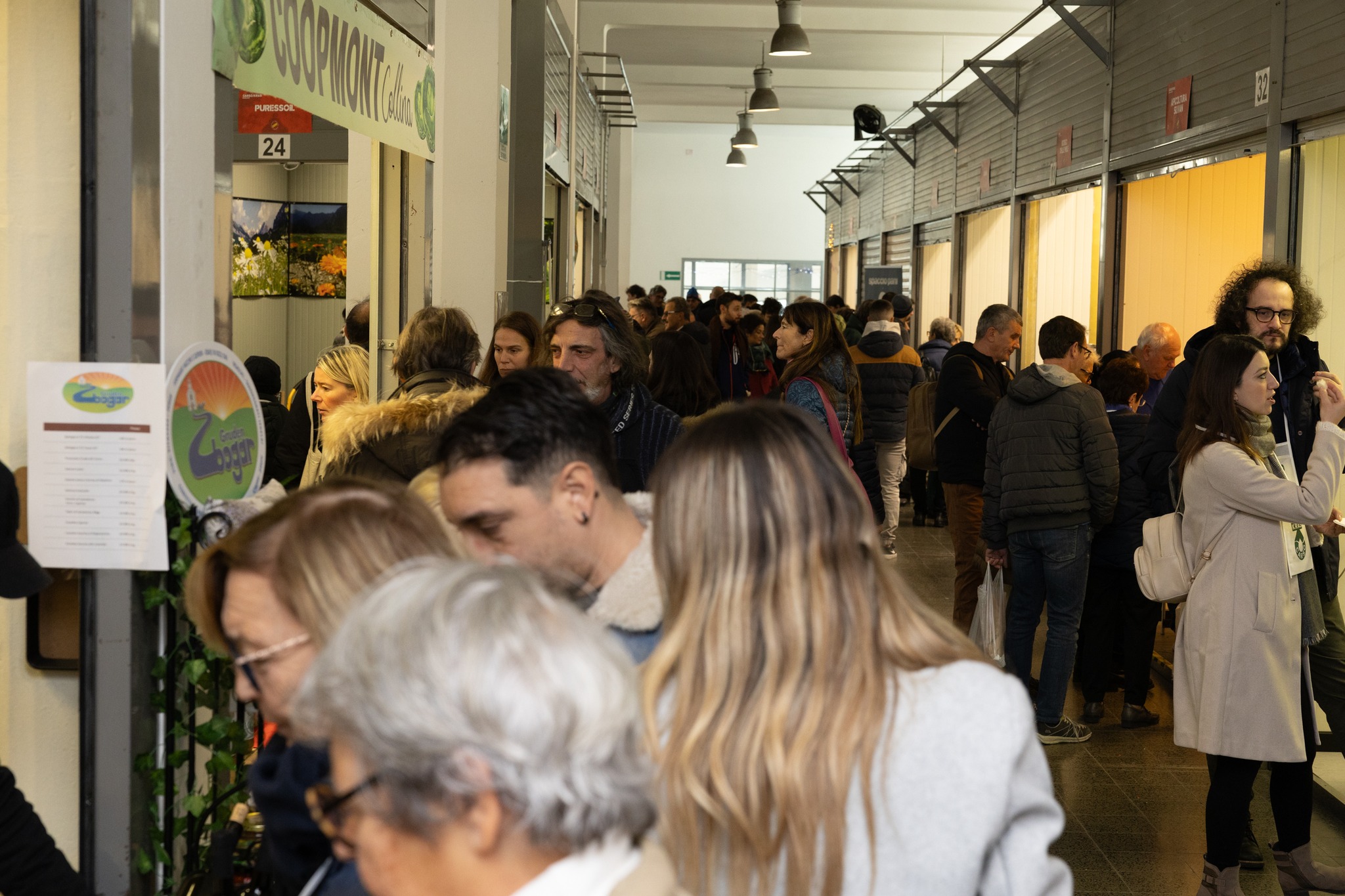 A bustling corridor inside the old farmers' market building in Trieste, filled with visitors exploring various stalls showcasing local produce and artisanal products during the Farmer & Artist event. Vibrant displays and signs from vendors