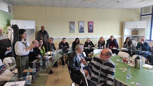 A woman talking to an audience in a room of people sitting around a table and listening