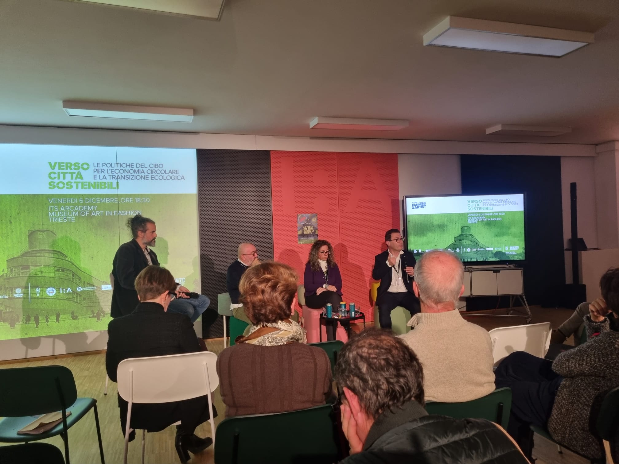 A panel discussion. Four speakers are seated on a stage in front of an audience. A projected presentation titled "Verso Città Sostenibili" is displayed on the wall behind them.