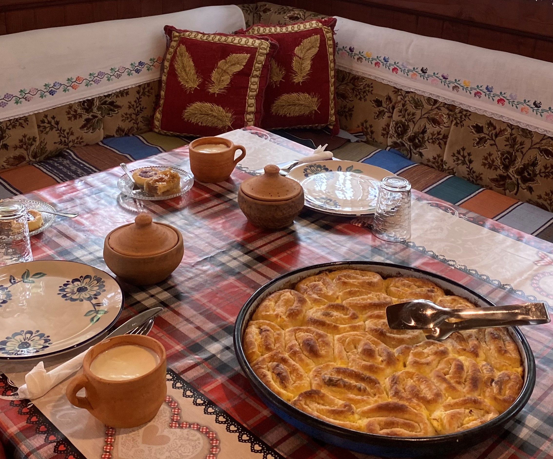 A cozy dining setup in a traditional Bosnian room featuring embroidered cushions, and a plaid tablecloth. The table is adorned with a freshly baked Bosnian pie in a large round dish, surrounded by rustic bowls, plates, and a cup. The warm and inviting ambiance reflects the region's rich cultural heritage.